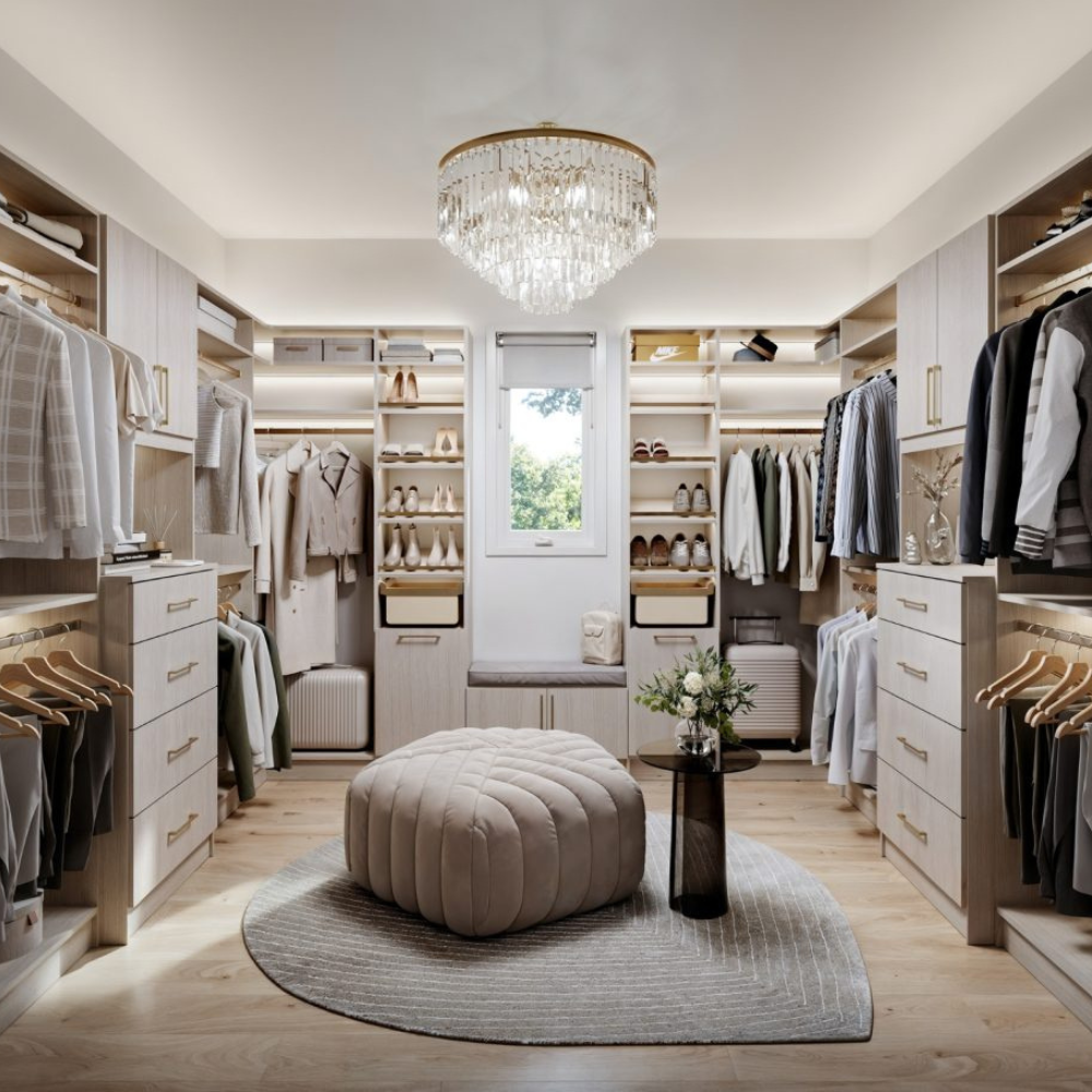 Luxurious walk-in closet with light wood shelving, hanging rods, a central tufted ottoman, and a crystal chandelier, featuring neatly organized clothes, shoes, and accessories.